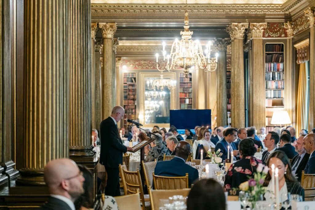 Man giving a speech at a formal dinner, guests seated at tables in a grand hall. - International Awards